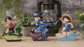 Three One Piece figures laid out on a wooden table, with plants and an old-fashioned lantern in the background